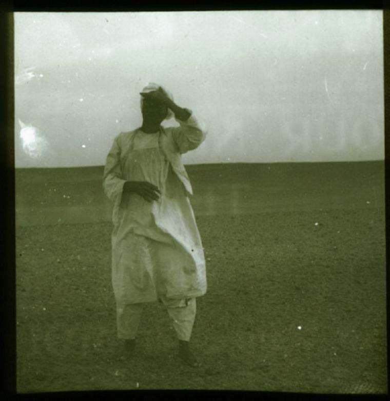 A man in white clothing stands alone in the foreground, an empty landscape behind him. Historical black & white photo.