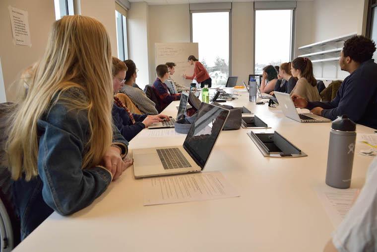 Students sitting around large desk watching professor write notes in front of class. Photo.