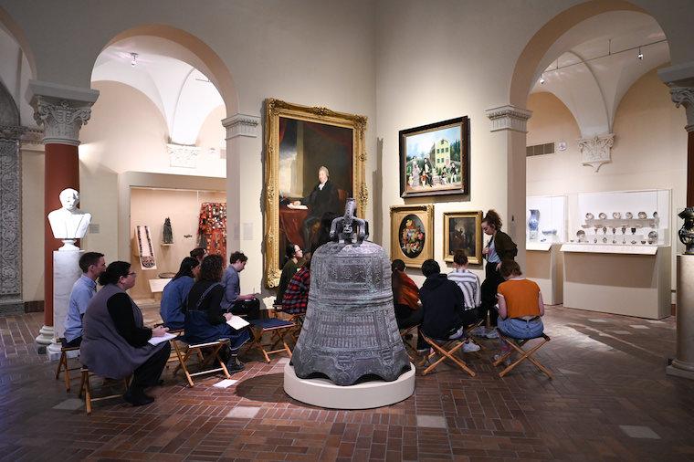 Students look at paintings in the museum while seated in chairs in the gallery.