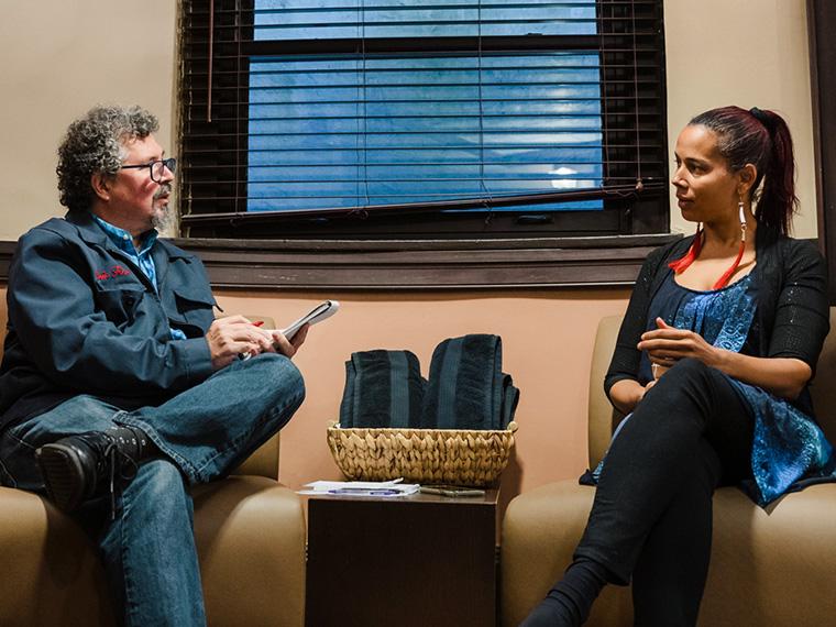 Seated in comfortable chairs, Rhiannon and a writer speak. The writer is taking notes.