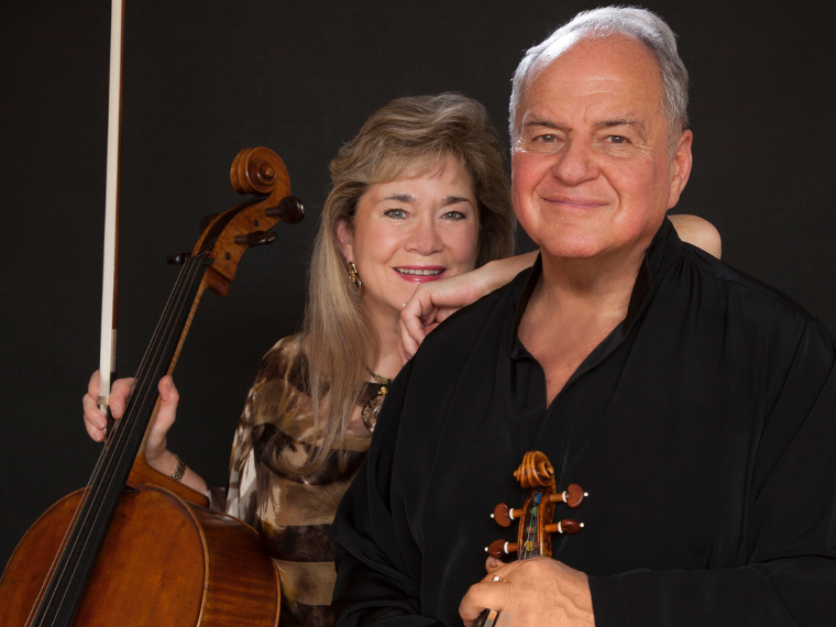 Man holding violin and woman holdiing cello and bow smiling at camera