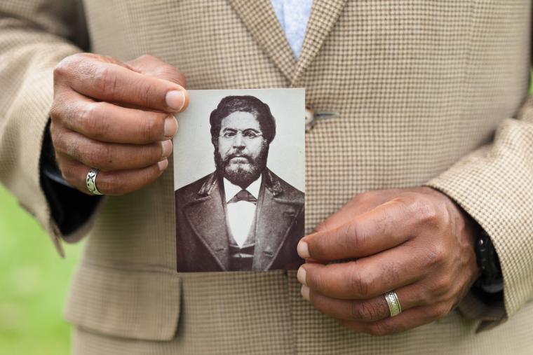 Hands holding an antique photo of Vashon shows a serious, bearded man with wire-rim glasses.