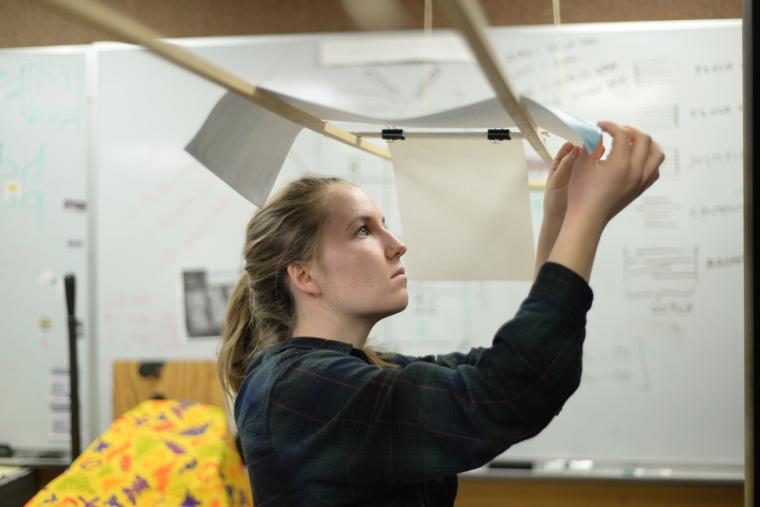 A student inspects a sheet of paper that is draped over 2 wooden rods above her..