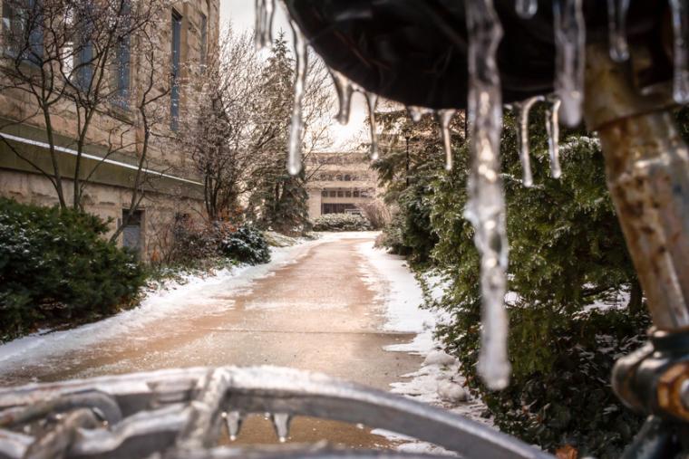 An icicle hangs from a bike seat in the foreground. Behind, a snowy path leads to a building.