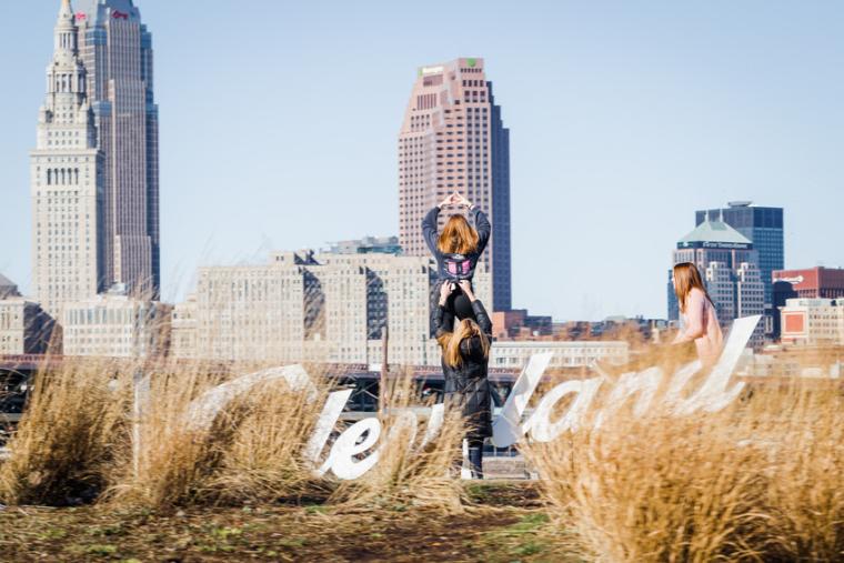 Photograph of a city backdrop, a sign that says, "Cleveland," and three women posing by the sign.