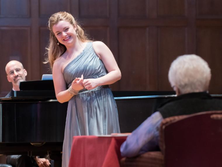A singer, standing by a grand piano, awaits feedback from Marilyn Horne, who is seated at a small table.