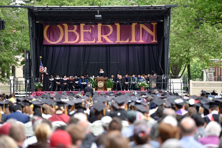 A view of the Commencement speakers on stage under an 'Oberlin' banner. The audience in the foreground is a sea of graduation caps.