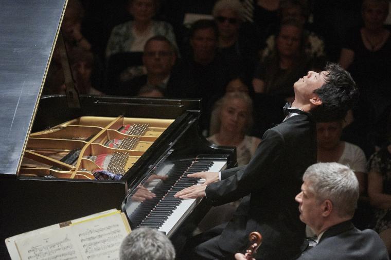 A pianist performs before an audience. He is leaning back with eyes closed and fingers on the keys.