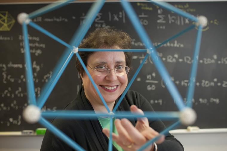 Susan Colley holding a geometric model. In the background, a chalkboard with equations.