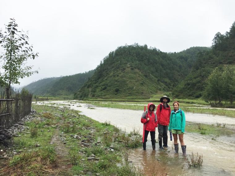 3 people in boots and rain gear stand in a shallow, muddy river.