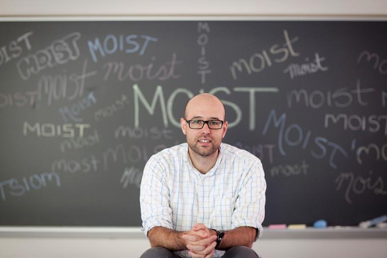 Man seated in front of chalkboard showing the word 'moist'