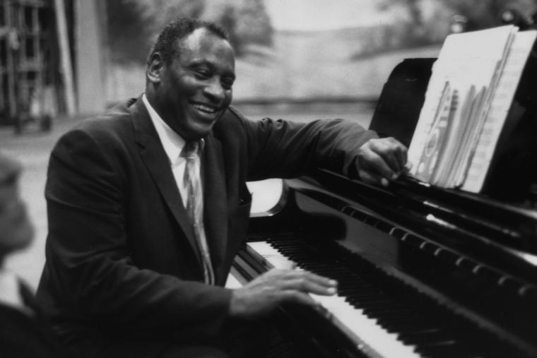Paul Robeson seated at a piano, sharing a laugh with someone. Black and white photo.