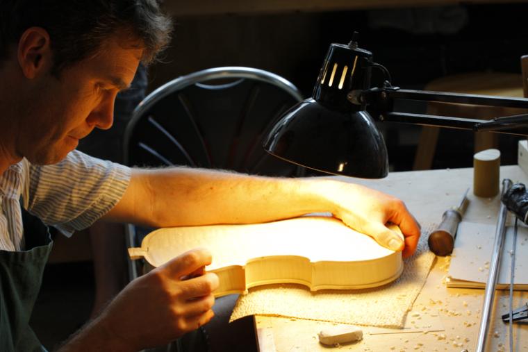 Under a bright lamp, a man works on unfinished wood in the shape of a violin body