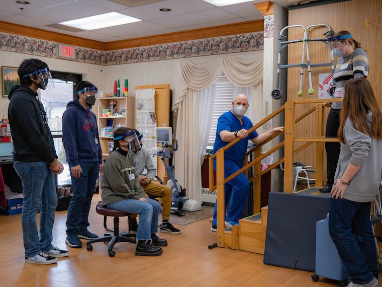 Six students surround nursing home attendant who demonstrates how to help patients up steps