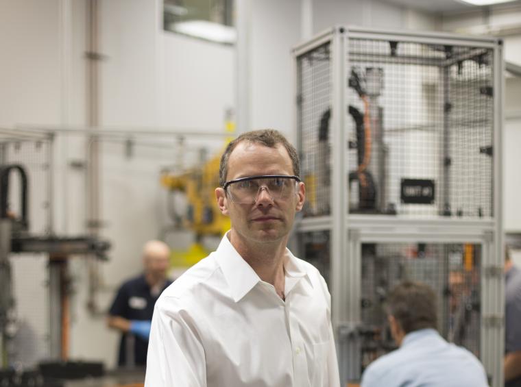 A man in a large workshop wears safety goggles and a white dress shirt. Behind him are two workers and a metal cage containing an apparatus. 
