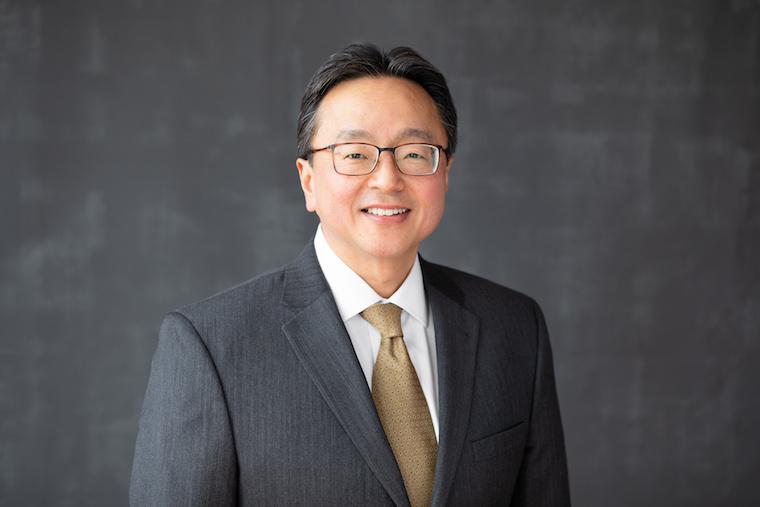 David Kamitsuka smiling with glasses gray suit coat and tan tie.
