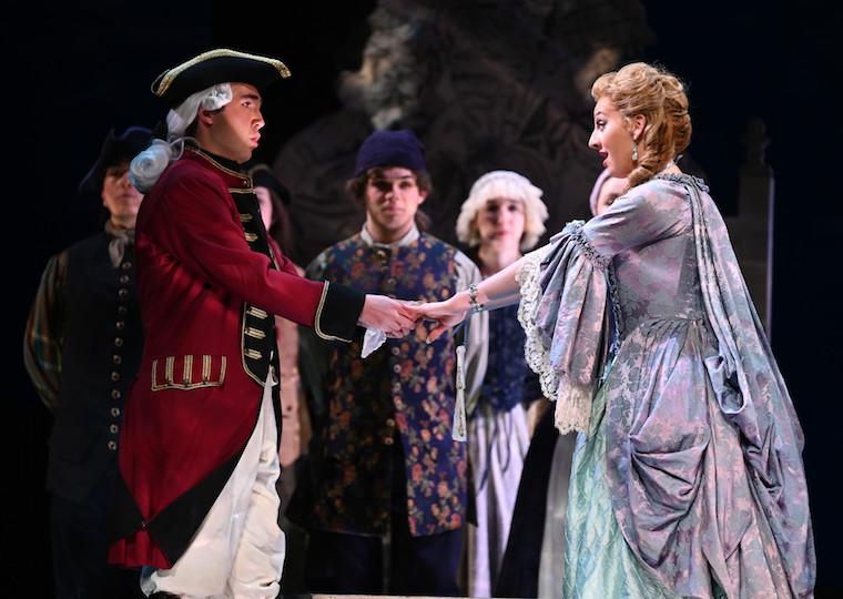 Two actors dressed in 18th century costume hold hands on stage.