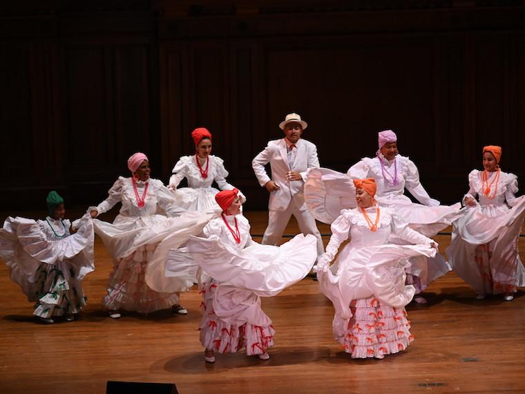 Seven women dance in full skirts and a man dances with them on stage.