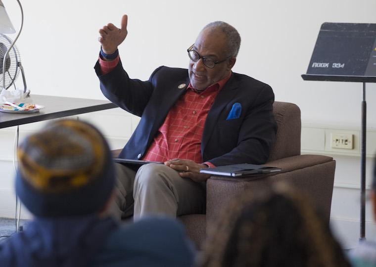 A man sits in front of a group of students, holding his hand out.