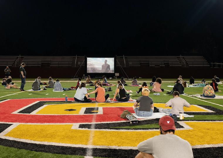 Students sit on a football field and watch a movie on a large screen.