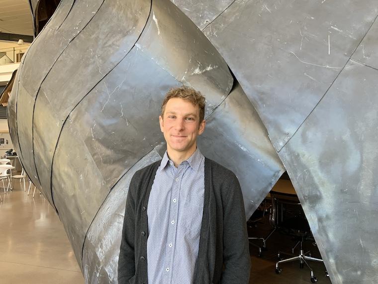 Young man in blue denim shirt nd dark gray sweater standing in front of metal sculpture.