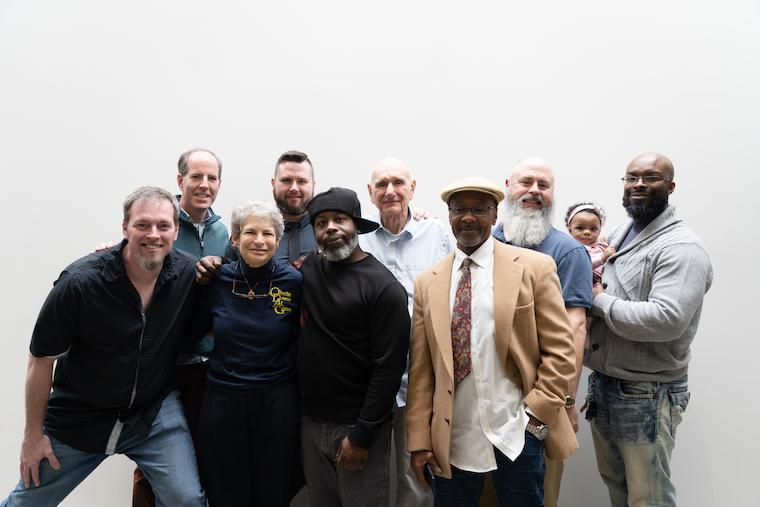 Eight white and black men of all ages, one woman, and a man holding a baby post for photo.