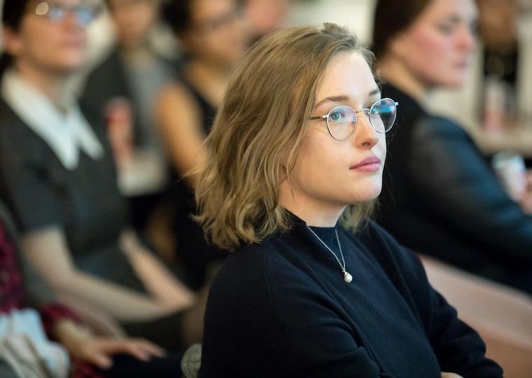 A girl wearing glasses sitting in an audience.