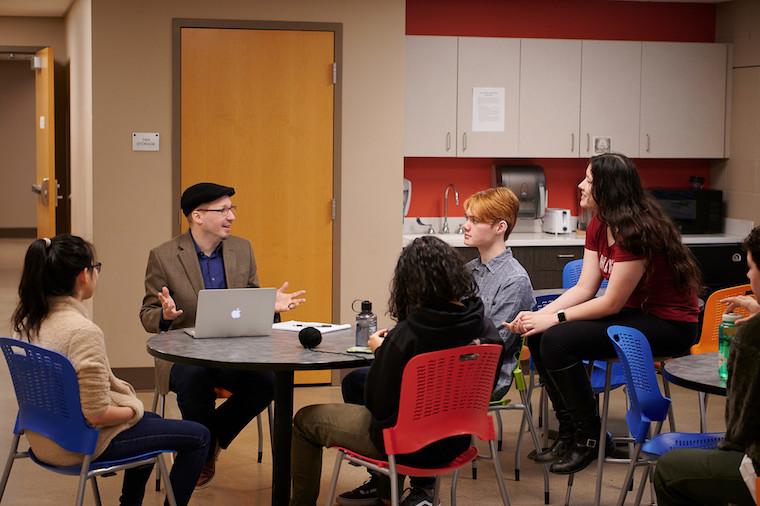 Four students and professor talk while seated at a round table.