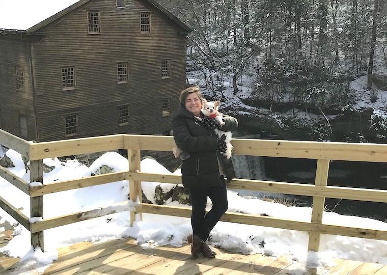 A woman holds a small dog while standing on a wooden ramp near a barn.