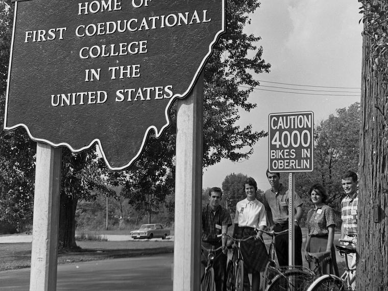 A small group of students stand next to a bicycle sign while holding their bikes.