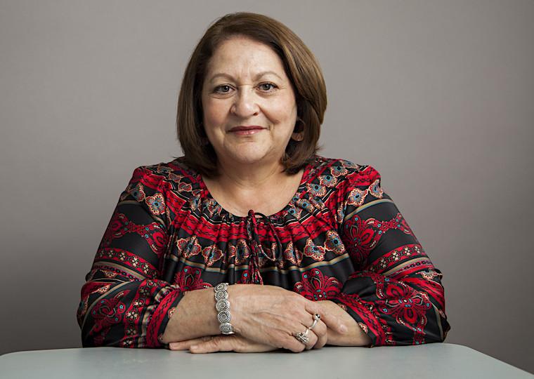 A portrait of a middle-aged woman wearing with arms resting on a table.