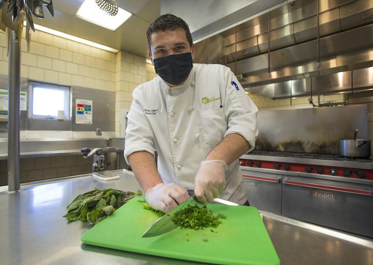 A man wearing a chef jacket chops parsley on a cutting board.