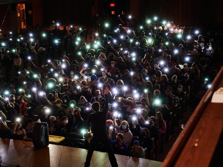 A large audience hold up cell phones in a dark concert hall.