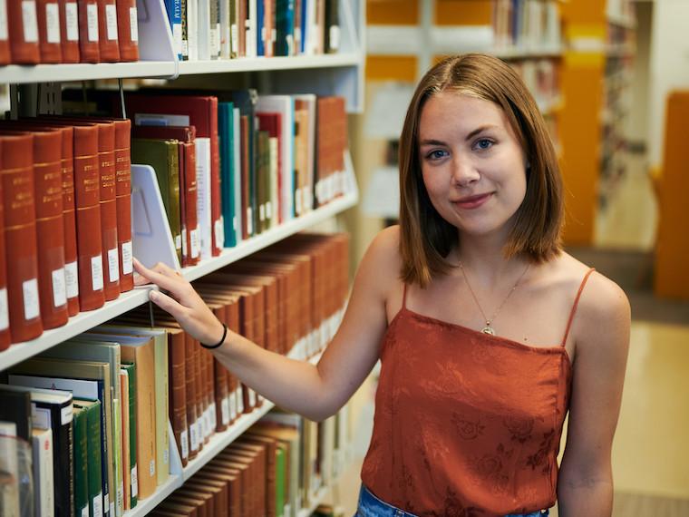 A portrait of a college student standing next to a library bookshelf.