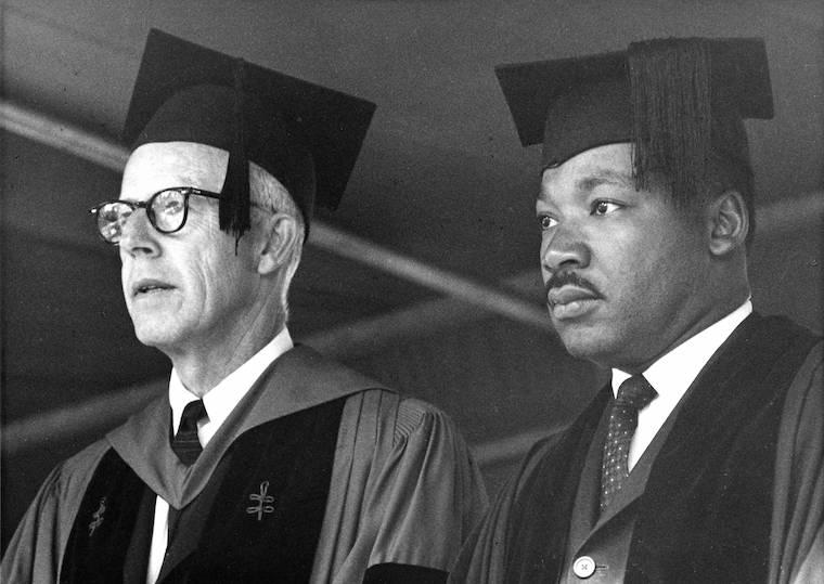 Two men wearing graduation caps and robes look out into a crowd.
