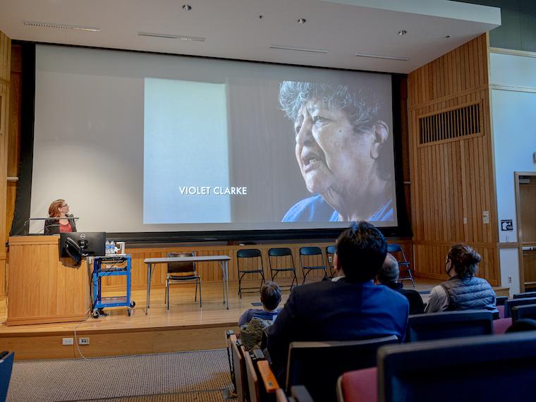An elderly woman's face is shown on a large projector screen.