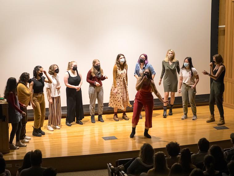 A semicircle of students stand around a student in the middle.