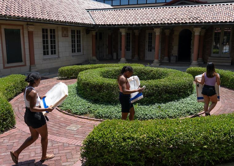 Three students walk through a courtyard while holding pieces of artwork.