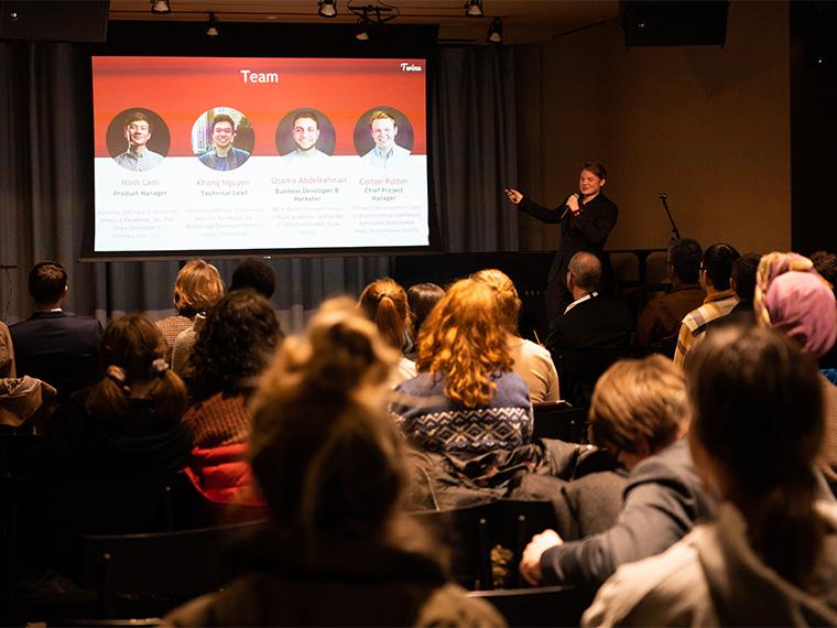 A large screen with the faces of the Twine app team, while an audiences watches.
