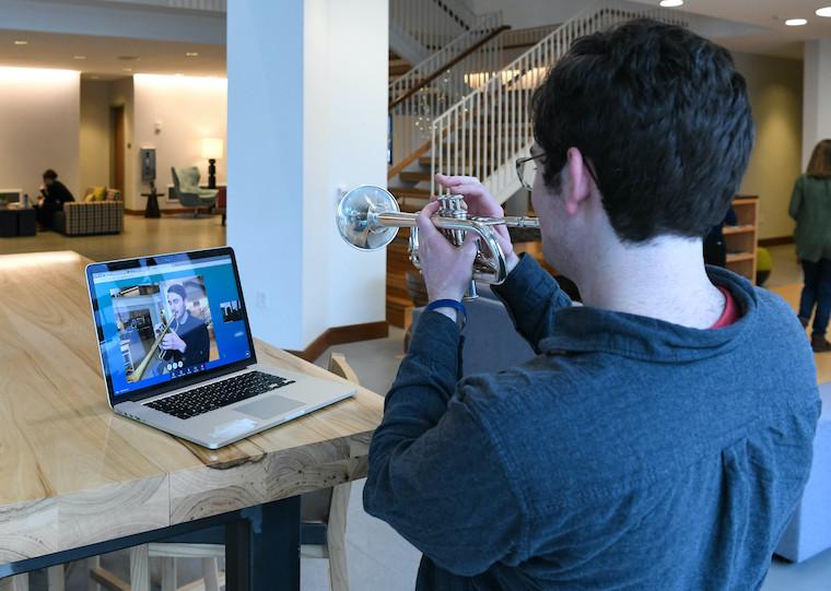 A college student plays a trumpet while a person on a computer screen facing him plays a trumpet.