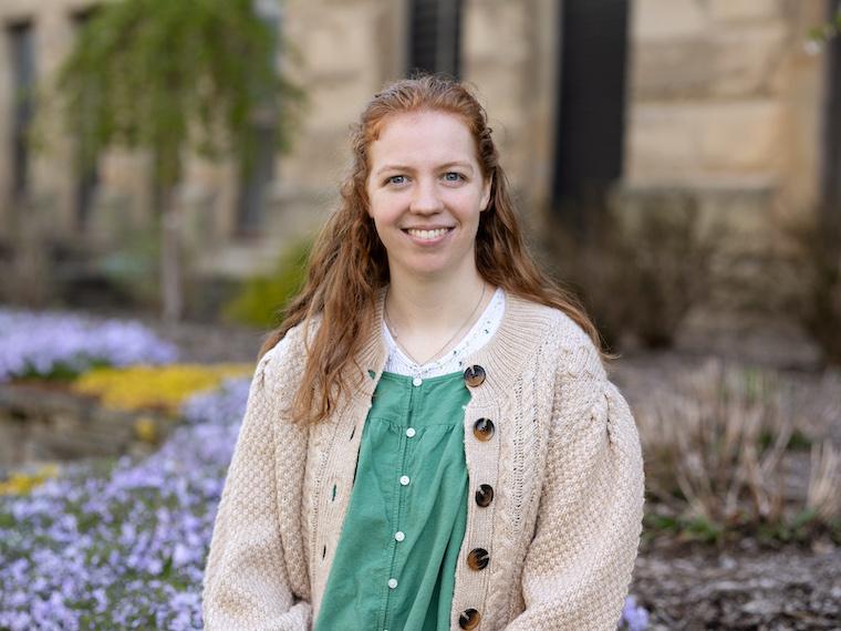 A portrait of a female college student sitting next to flowers on a brick wall.