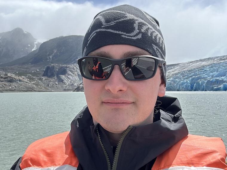 A man wearing a sweater cap and jacket stands in front of mountains and water