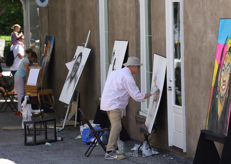A row of artists draw on canvases in a small alley.