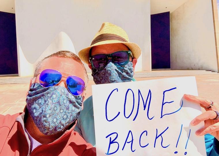 Two men wearing masks and sunglasses hold a sign that says come back.