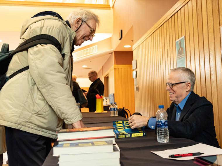 A man signs a book at a book signing for another man standing in front of him.