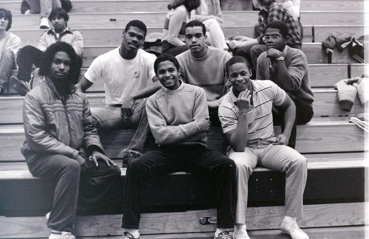 Six male students pose for a photo on gymnasium bleachers.