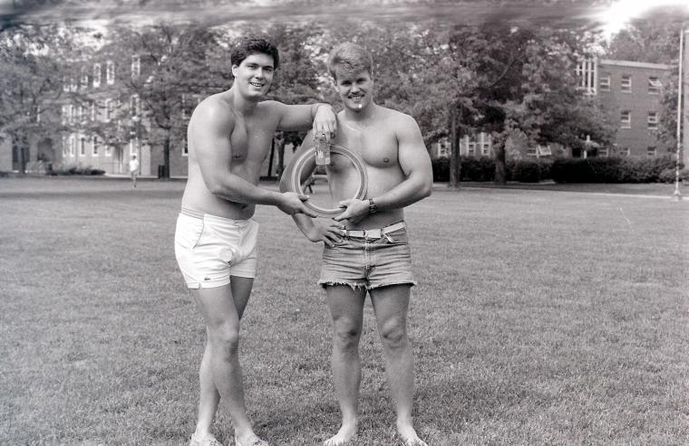 Two shirtless male students pose outside on campus.