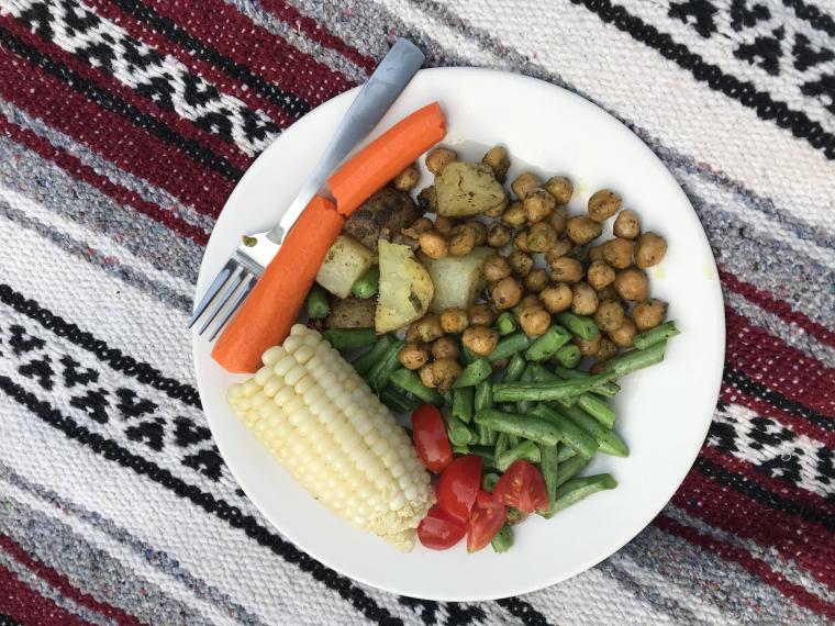 a plate of food with vegetables, corn, potatoes, and chickpeas on a picnic blanket