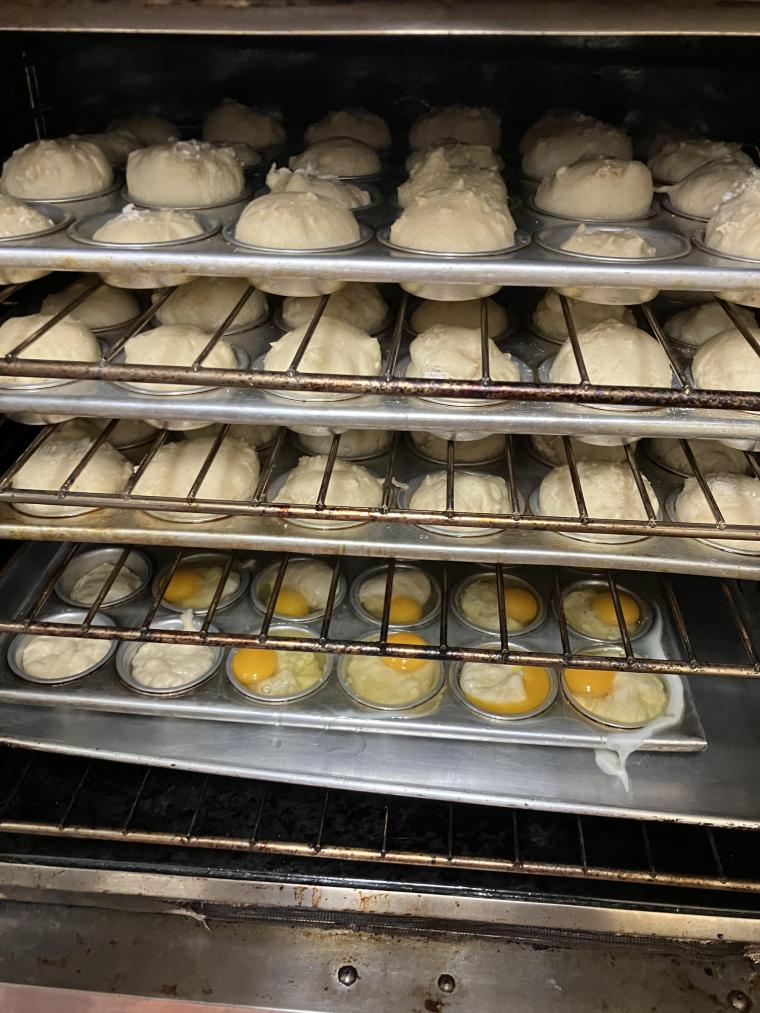 Picture of the inside of an oven. There are four racks. On the upper three racks there are rolls baking in muffin tins. On the lowest rack there are raw sunny side up eggs baking in muffin tins.