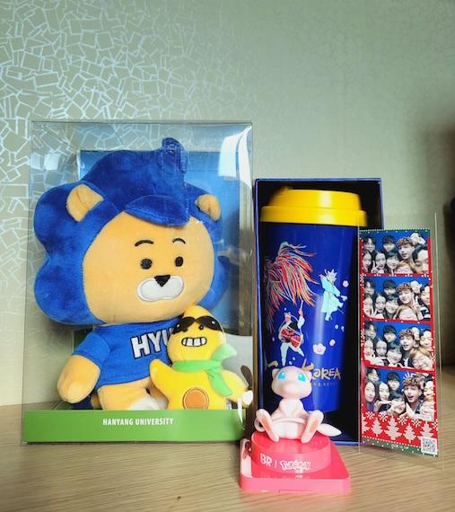 A plush doll of Hy-Lion in a plastic box, a limited edition "Korea" Starbucks tumbler, a Mew figurine, and a photo-card of my friends and I.